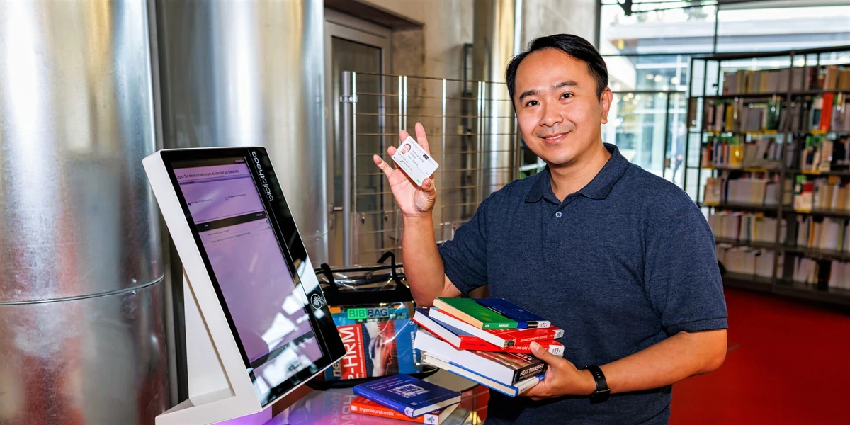  	Ein Student steht mit ein paar Büchern in der Hand am Ausleihautomat und hält in der anderen Hand seinen Studentenausweis hoch.