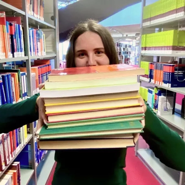 Eine Studentin in der Hochschulbibliothek mit einem Bücherstapel in beiden Händen