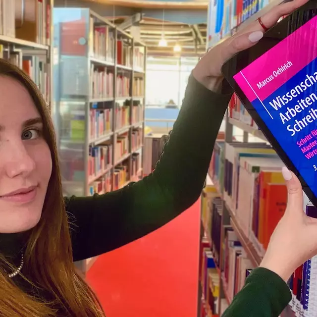 Studentin zieht ein iPad mit geöffnetem E-Book aus einem Bücherregal