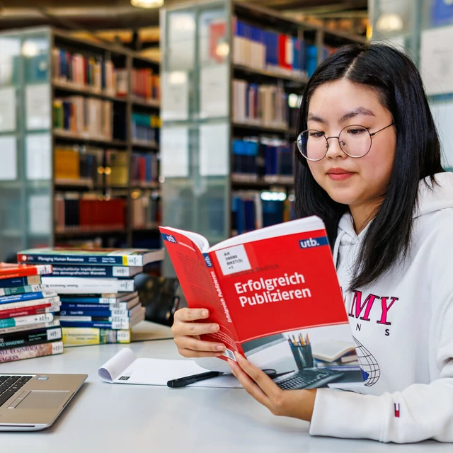 Eine Studentin sitzt im Lesesaal an einem Arbeitsplatz mit einem Buch mit dem Titel Erfolgreich Publizieren in Händen. Am Arbeitsplatz befindet sich ein Laptop auf dem die Startseite des Publikationsservers der OTH Regensburg zu sehen ist.