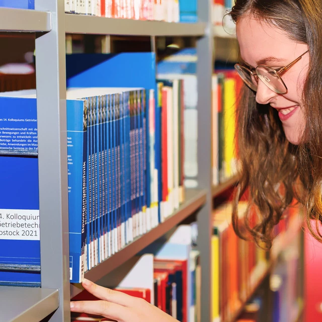 Eine Studentin sucht am Buchregal nach einem Buch und deutet auf die Signatur.