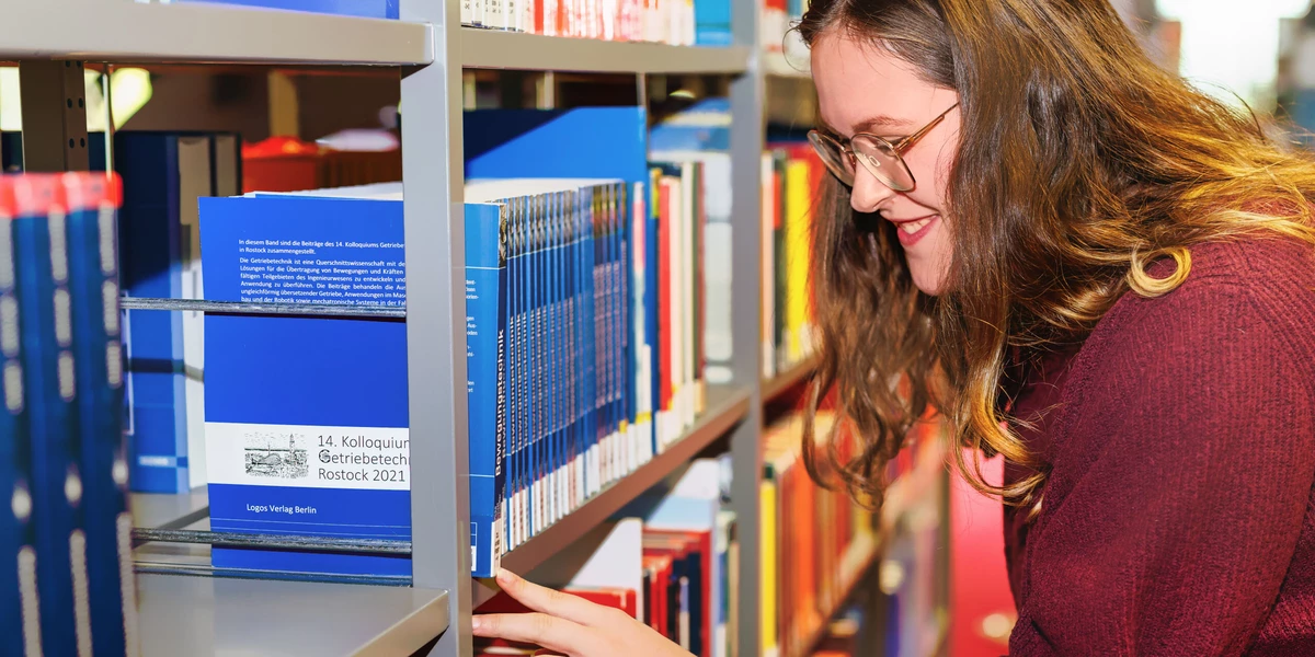 Eine Studentin sucht am Buchregal nach einem Buch und deutet auf die Signatur.
