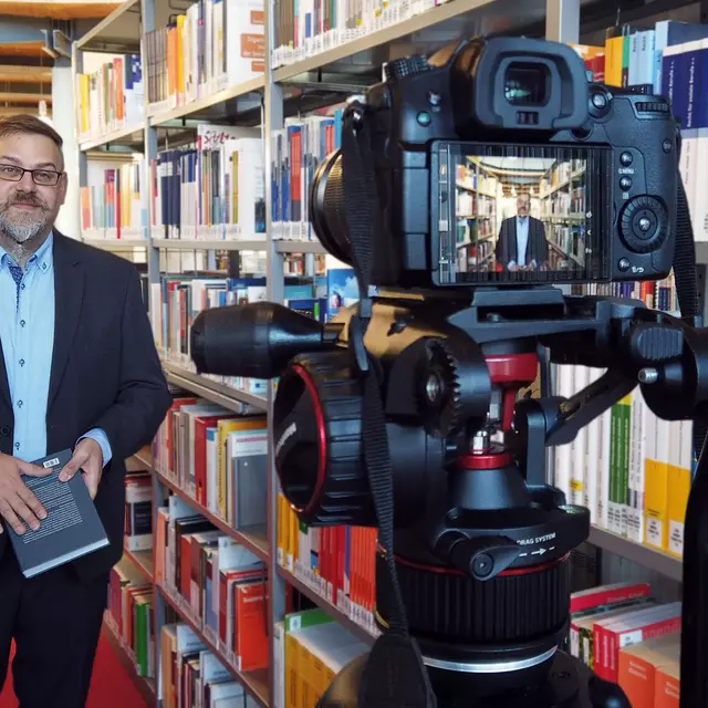 Ein Bibliotheksangestellter steht im Lesesaal zwischen Bücherregalen vor einer Videokamera mit einem Buch in der Hand.