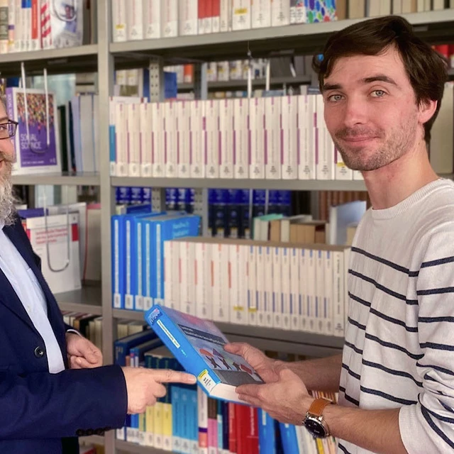 Ein Bibliotheksangestellter deutet auf die Signatur eines Buches, das ein Student in der Hand hält.