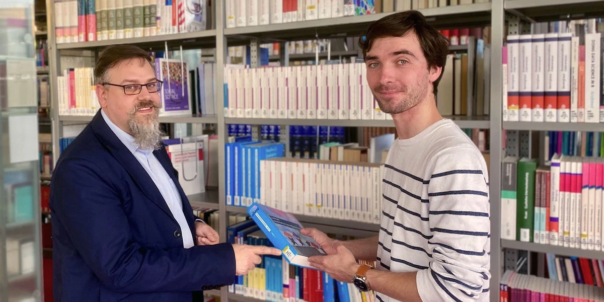 Ein Bibliotheksangestellter deutet auf die Signatur eines Buches, das ein Student in der Hand hält.